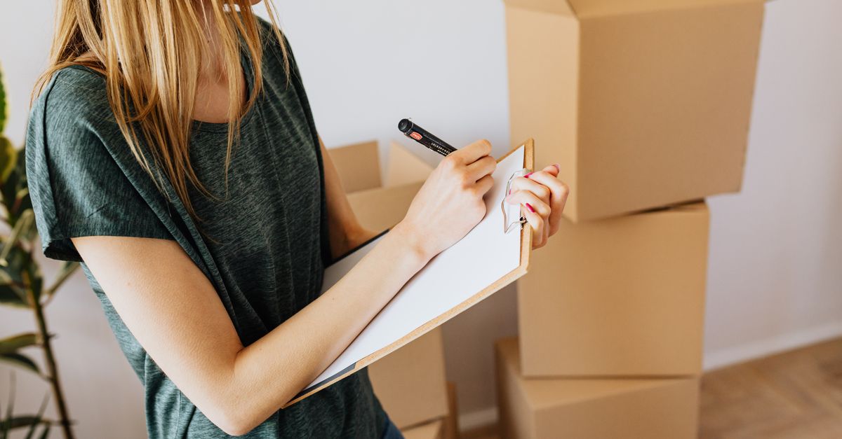 Side view crop female wearing casual shirt standing near packed cardboard containers and taking notes before relocation into new modern place